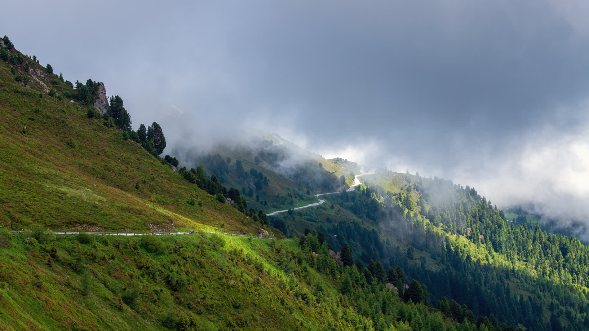Bergstraße am Hang, Wolken hängen tief über dem Wald, einzelne Sonnenflecken auf der Wiese