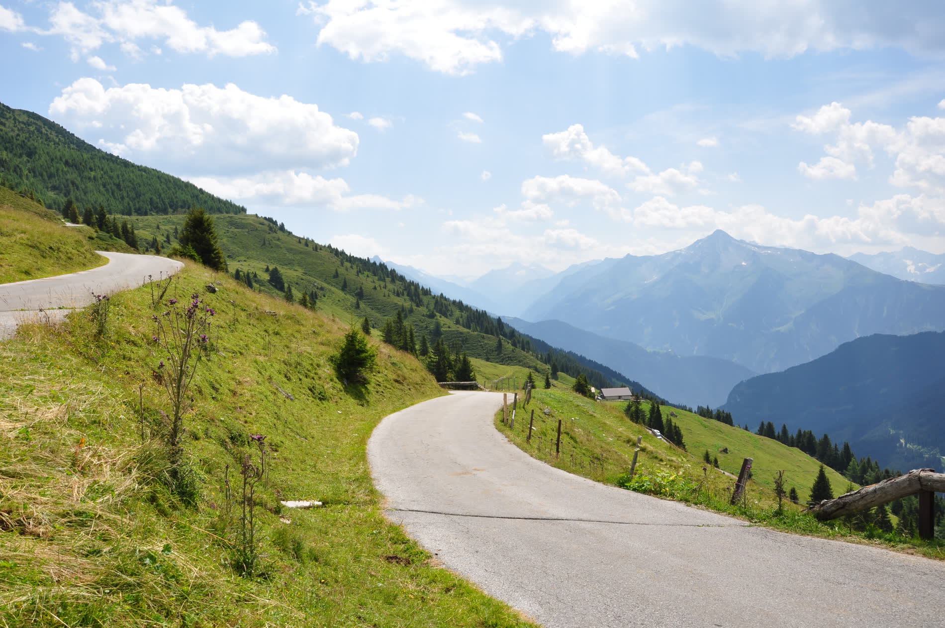 Schmale Asphaltstraße am grünen Almhang, rechts ein Holzzaun, dahinter Berggipfel im Dunst